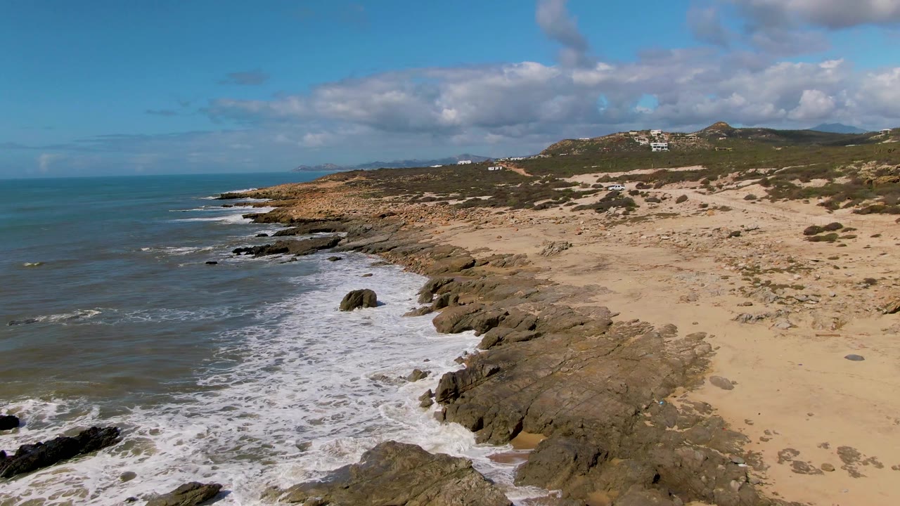 Aerial view at Buzzards Beach East Cape in Mexico with white foam spreading around nearshore rocks, open sand to the right, scrub vegetation beyond it, and no visible people.