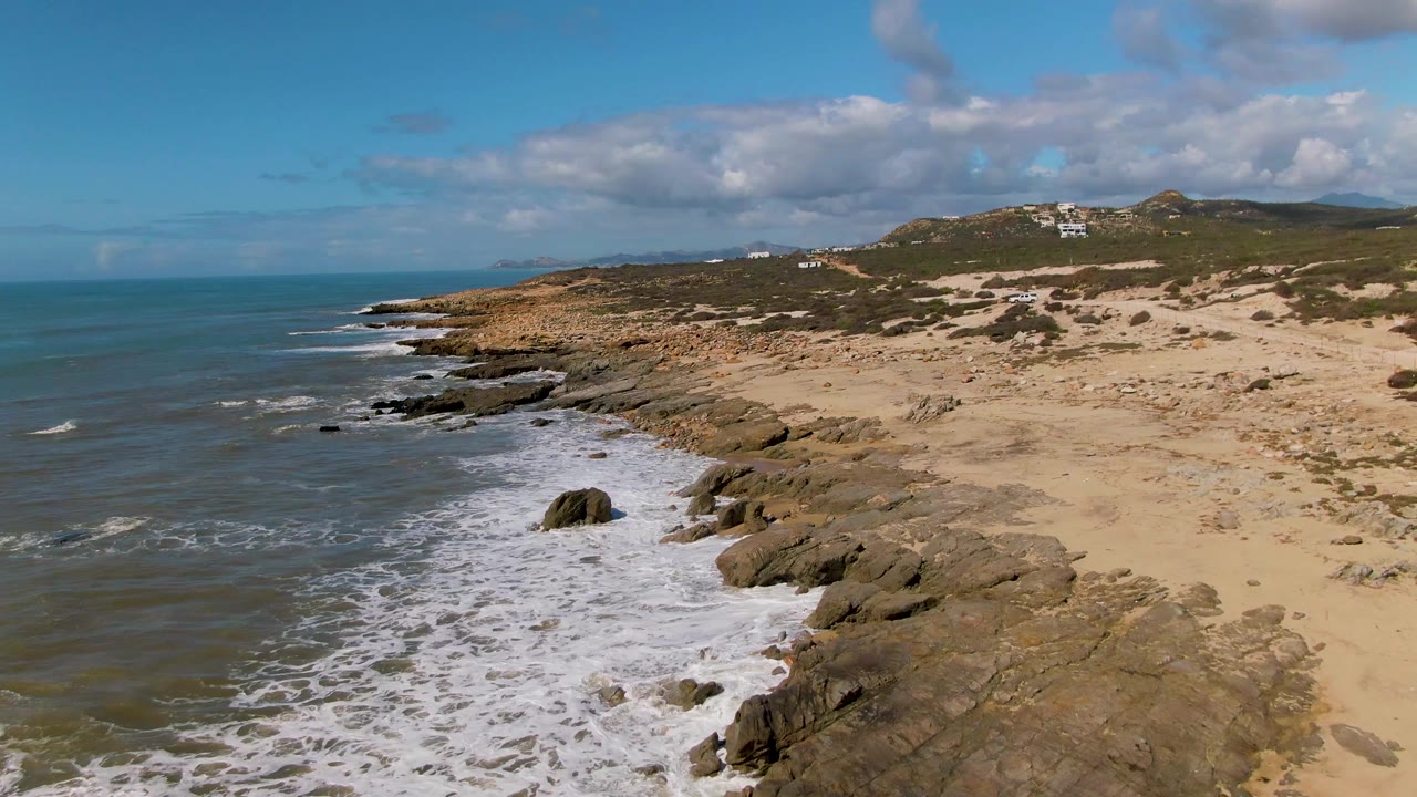 Aerial coastal frame from Buzzards Beach East Cape in Mexico showing a diagonal line of rocks, receding foam, broad tan sand, low green vegetation, and no visible people.