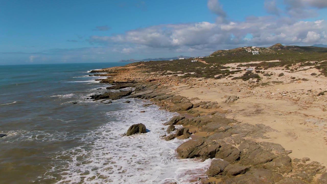 Aerial coastline with white surf, exposed rocks, dunes, and open ocean.