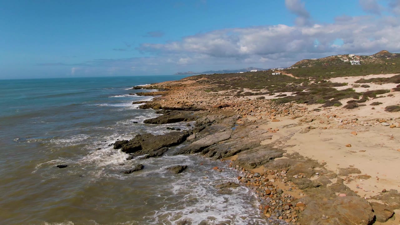 Waves curl into dark rocks along a sandy, scrub-covered coast.