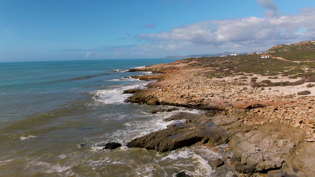 Aerial view at Buzzards Beach East Cape BCS Mexico showing green-blue water breaking against tan rock shelves, scrub-covered land, distant buildings, scattered clouds, and no visible people.