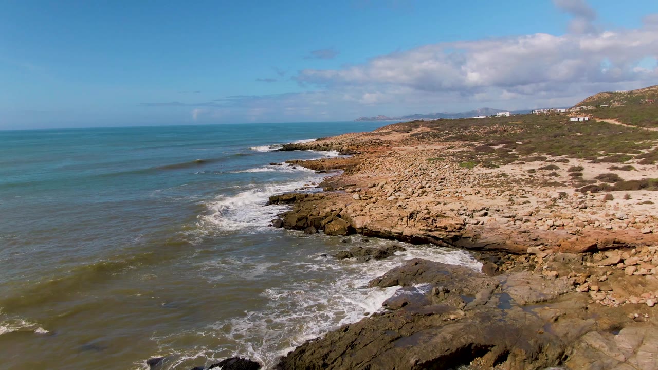 Aerial frame at Buzzards Beach East Cape BCS Mexico with white foam spreading over dark rocks in the foreground, open water to the left, low coastal hills to the right, and no visible people.