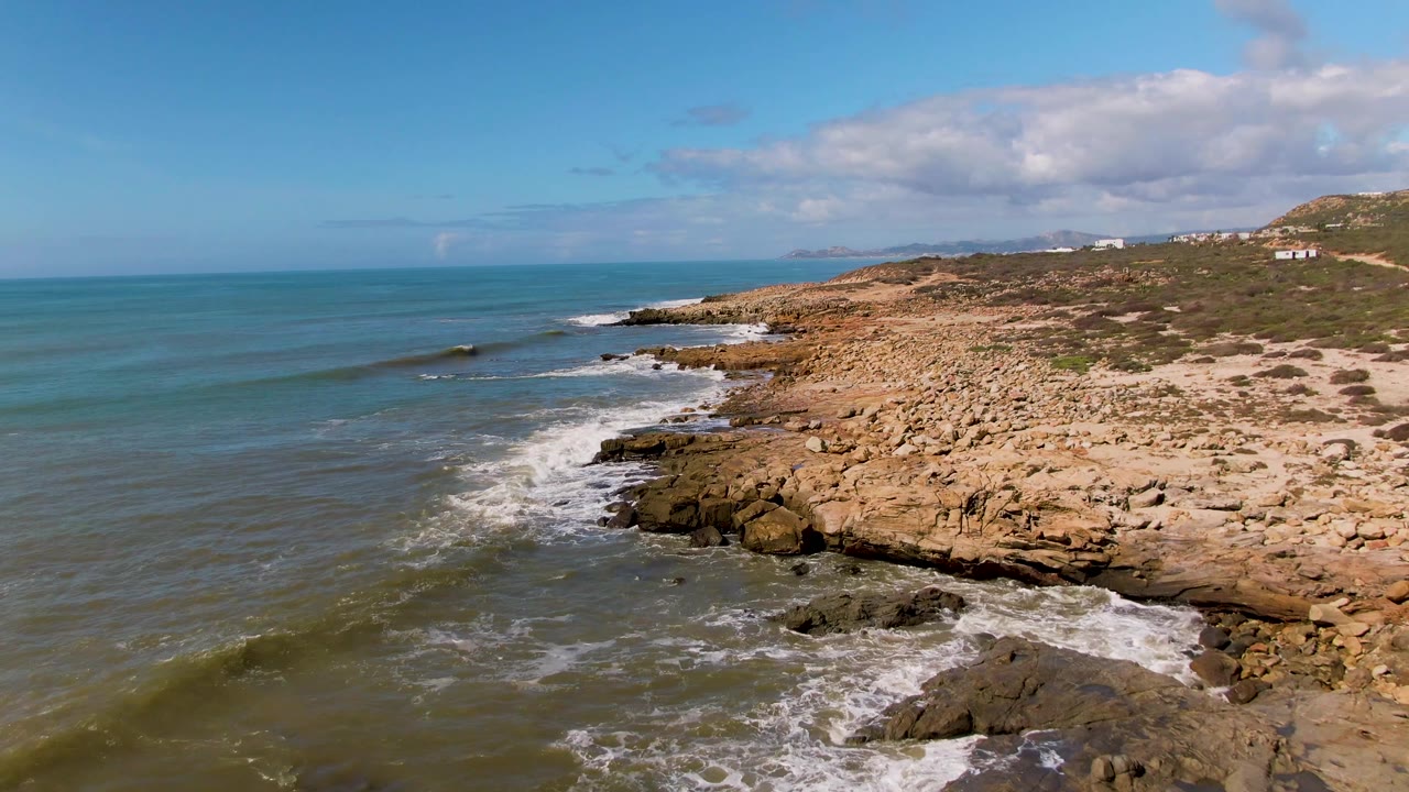 Aerial view at Buzzards Beach East Cape BCS Mexico showing a smaller wave breaking near offshore rocks, a long tan rock beach, greenish water, hillside buildings, and no visible people.