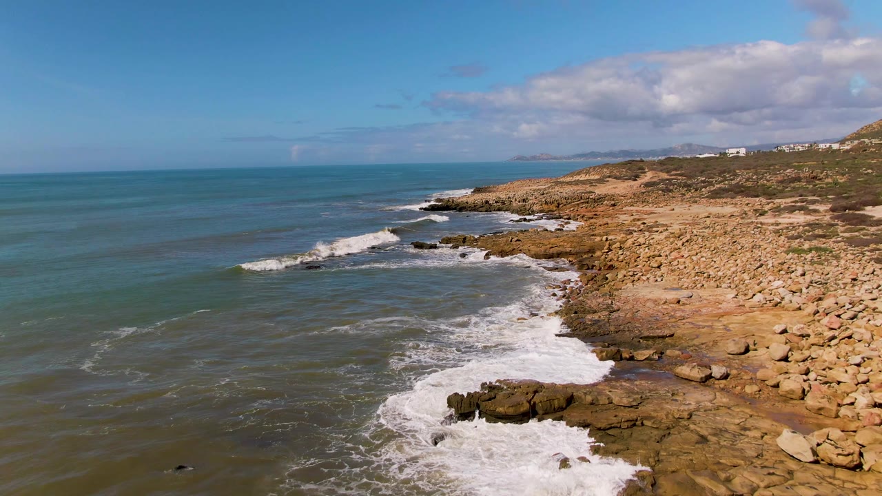 At 0:00, an aerial view over Buzzards Beach East Cape BCS Mexico shows blue-green ocean meeting a tan rocky shoreline, with white surf at the lower right and no visible people.