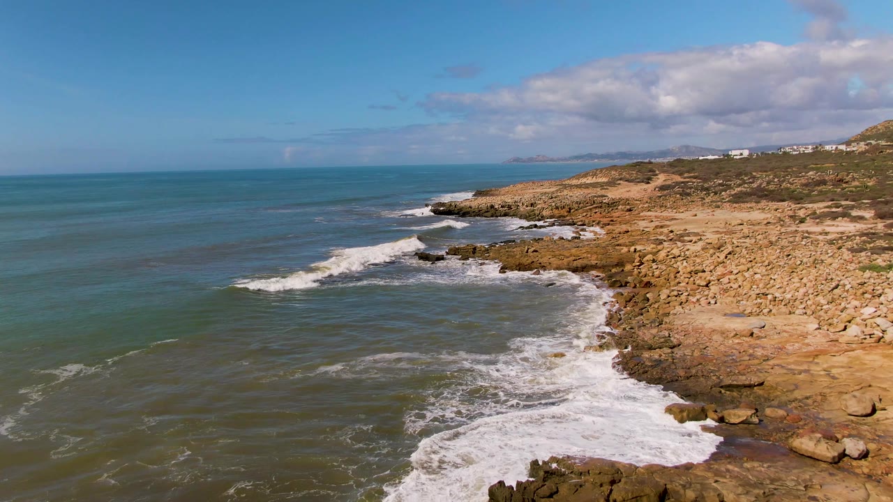Surf rolls toward tan coastal rocks with scattered clouds above the ocean.
