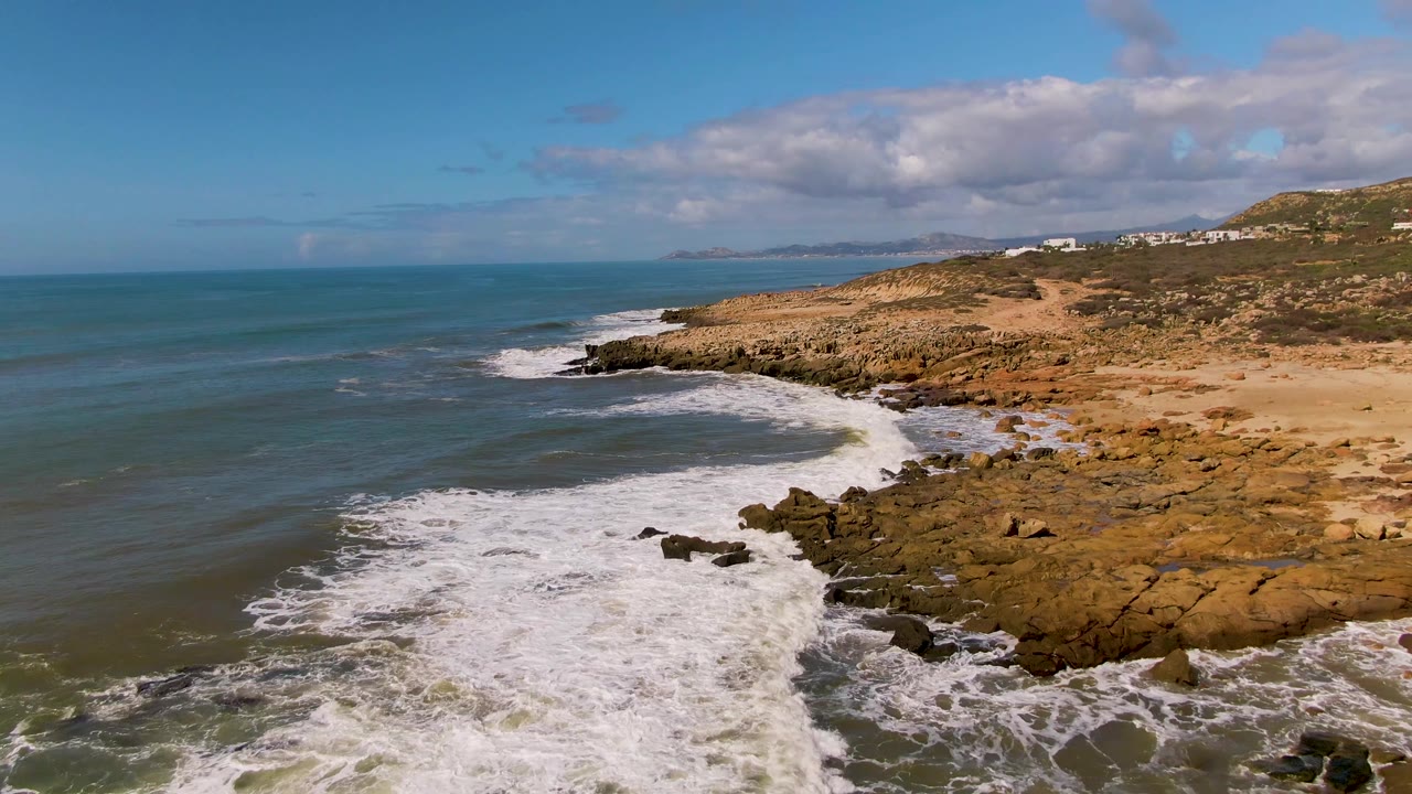 Aerial view at Buzzards Beach East Cape BCS Mexico with white surf spreading over dark shoreline rocks, blue-green ocean at left, tan coastal terrain at right, and no visible people.