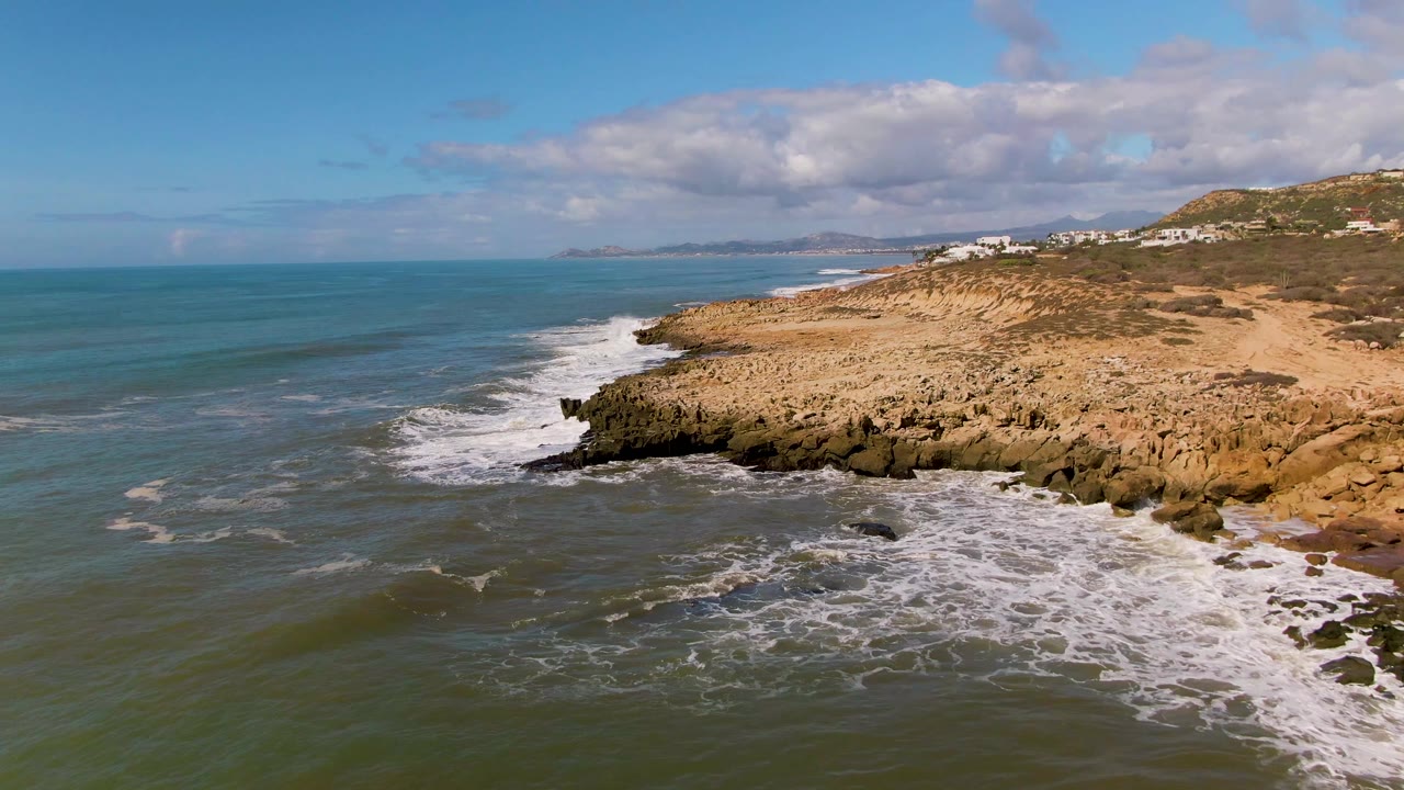Aerial frame over Buzzards Beach East Cape BCS Mexico showing green-blue ocean, white surf on dark rocks, sandy cliff top, distant hillside buildings, and no visible people.