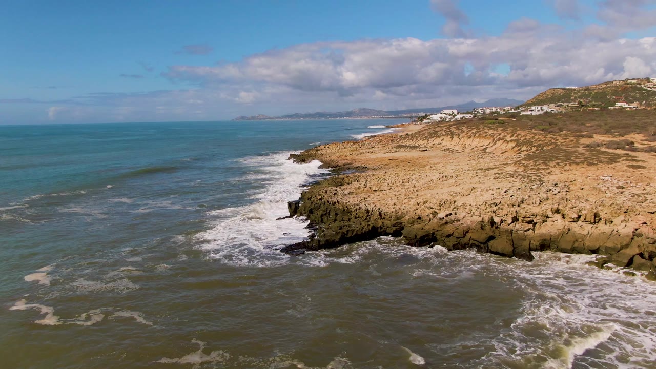 Buzzards Beach East Cape BCS Mexico coastline seen from a drone angle, with a breaking wave curling toward the rocks, sandy terrain on the right, distant buildings on the hill, and no visible people.