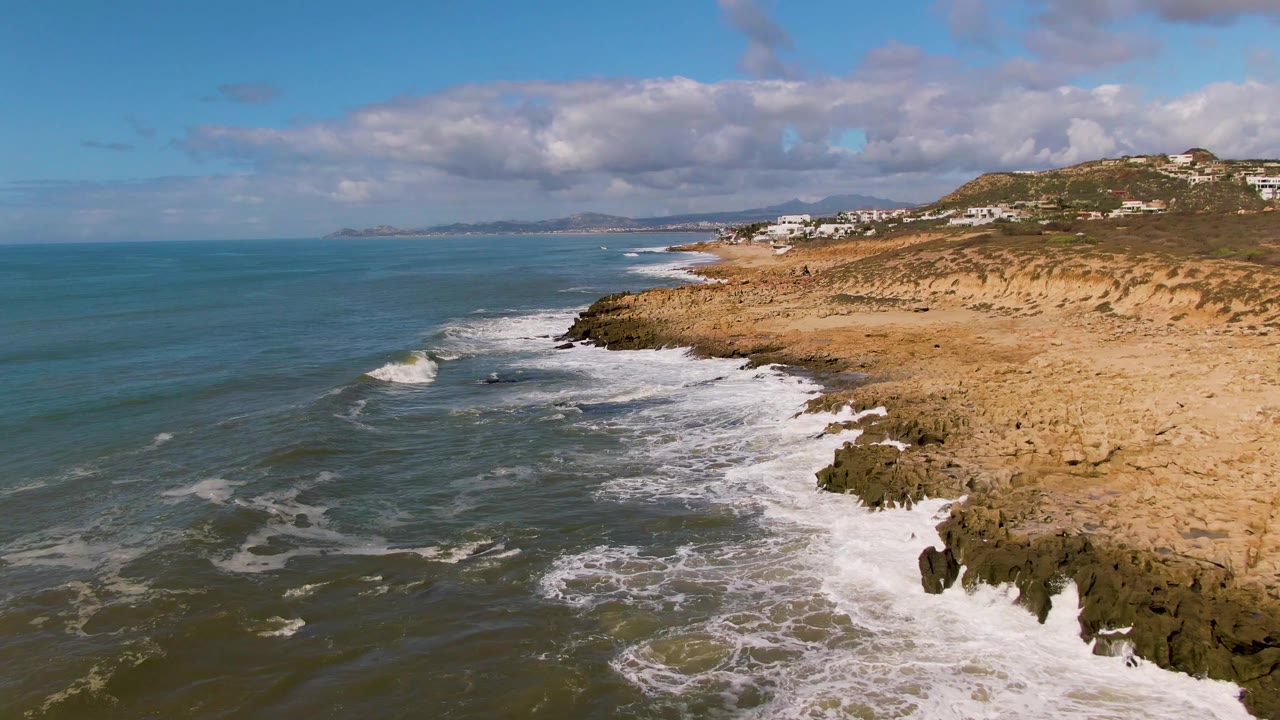 At Buzzards Beach East Cape BCS Mexico, white waves break along dark rocks below sandy cliffs, with blue-green ocean to the left and no visible people.
