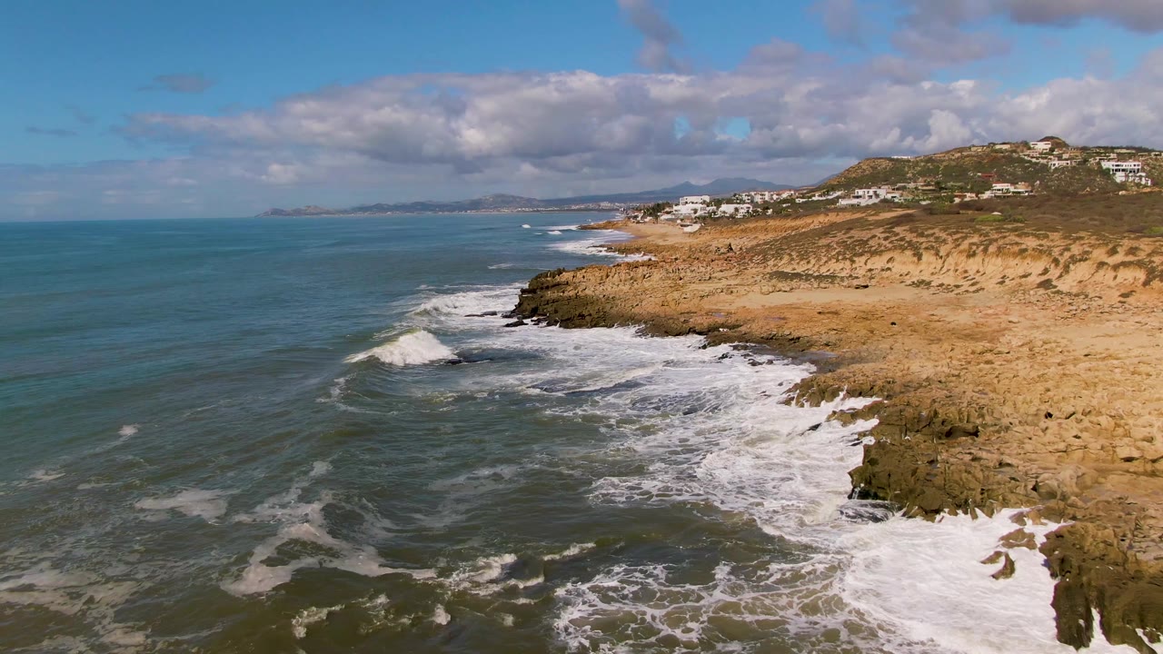 At Buzzards Beach East Cape BCS Mexico, a fresh band of foam pushes toward the rocky shore while distant white buildings line the hillside, with no visible people.