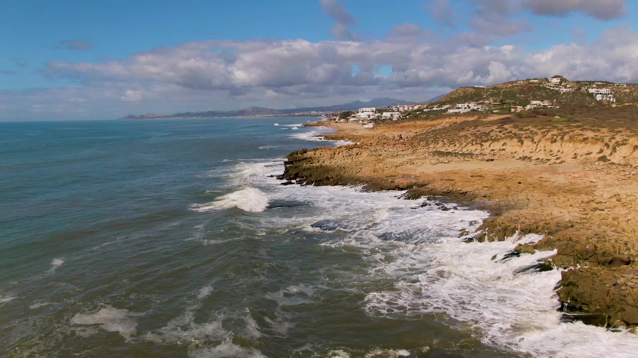 At Buzzards Beach East Cape BCS Mexico, white foam spreads wider across the rocks below the sandy bluff as the coastline recedes toward the horizon, with no visible people.
