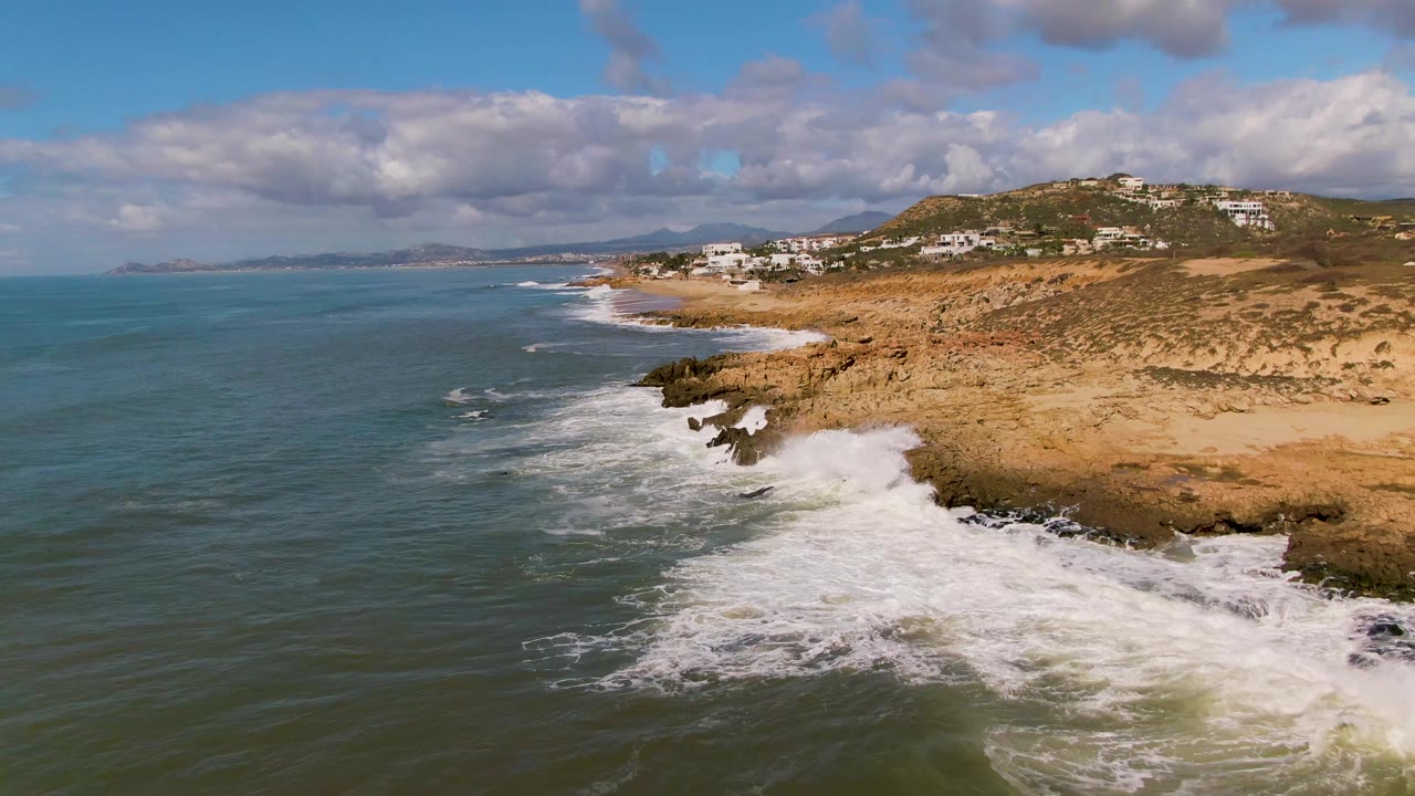 Aerial view at Buzzards Beach East Cape BCS Mexico with blue-green ocean on the left, white surf curling into dark rocks, tan coastal ground, distant hillside buildings, and no visible people.