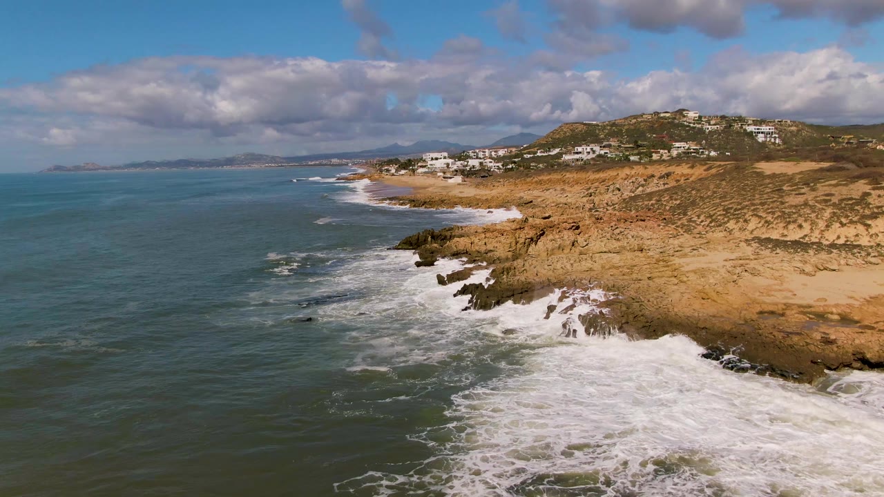White surf spreads over rocks beside a dry coastal hill and ocean.