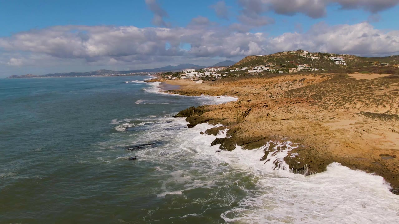 At 0:02, Buzzards Beach East Cape BCS Mexico is shown from above with a broader band of white foam along the lower right rocks, open water to the left, distant buildings on the hill, and no visible people.