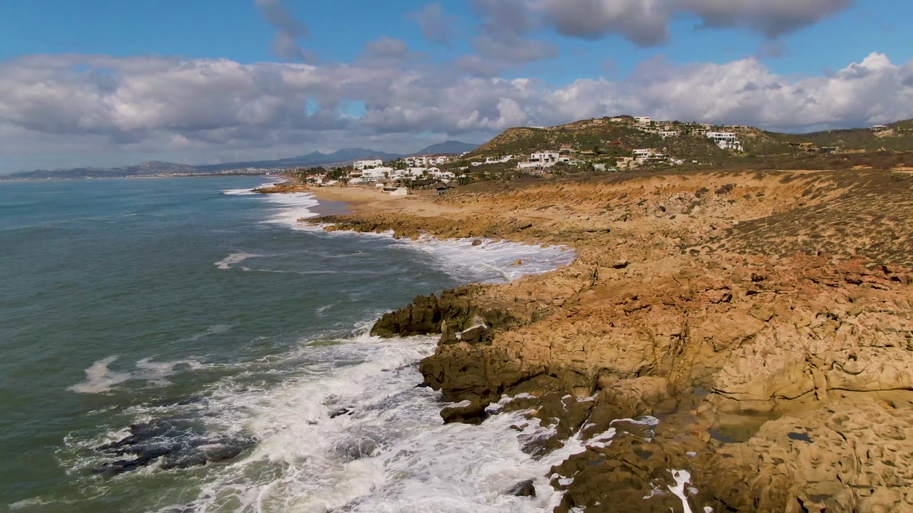 Opening frame of Buzzards Beach East Cape BCS Mexico, with teal ocean on the left, white surf along tan rocks, hillside buildings in the distance, and no visible people.