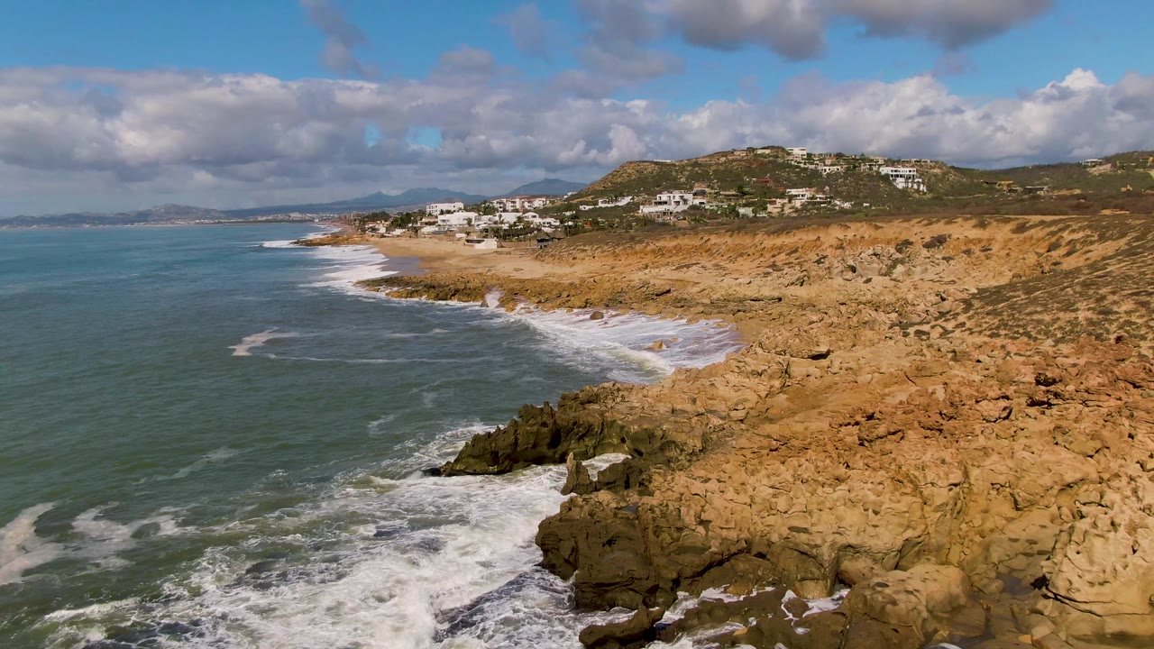 Foamy waves reach a jagged rock point along a tan coastal shore
