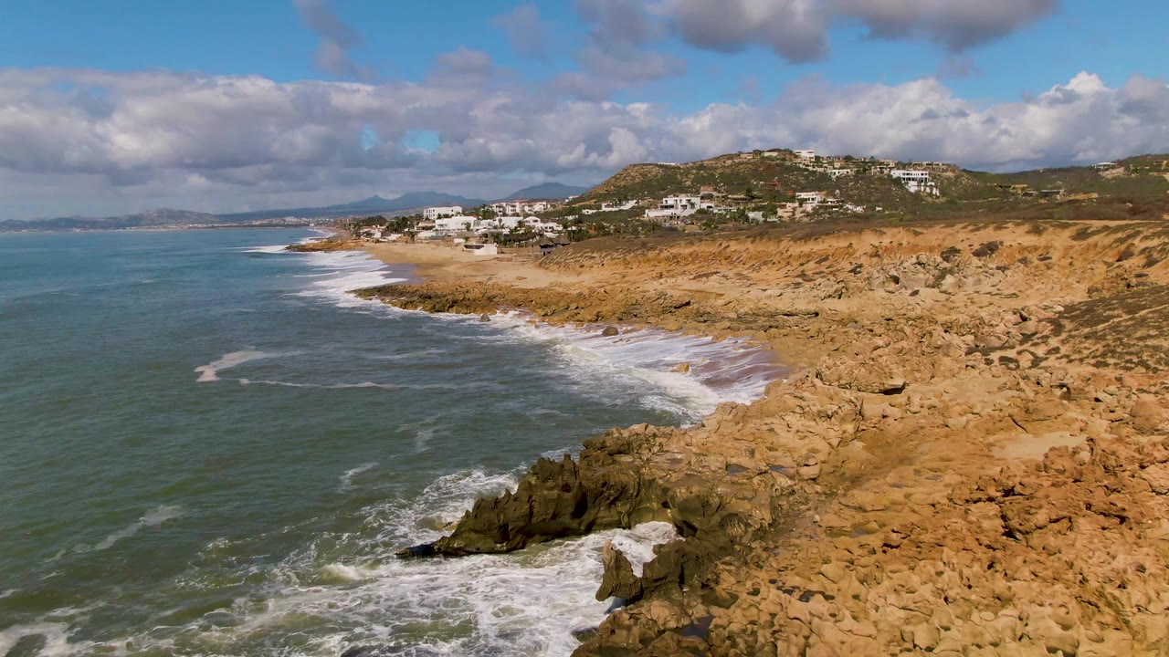 At 0:02, the aerial view shows broad surf bands sliding toward the rocky foreground at Buzzards Beach East Cape BCS Mexico, with houses on the hill and no visible people.