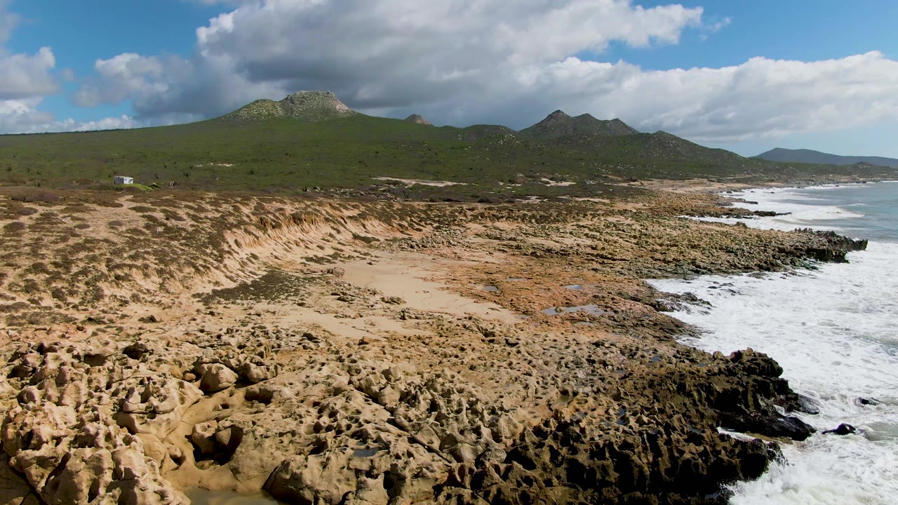 Aerial view of rocky dunes, surf, green mountains, and clouds