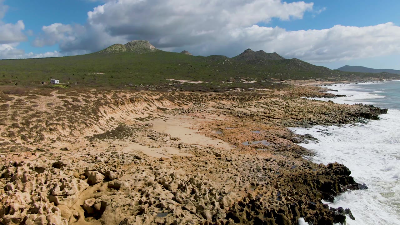 Aerial frame at 0:01 over Buzzards Beach East Cape BCS Mexico with more exposed rocky shore beside receding white water and no visible people.
