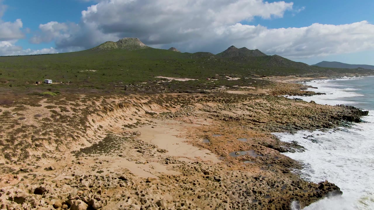 Aerial frame at 0:02 showing the Buzzards Beach rocky point, sandy dune patches, surf along the right side, green mountains, and no visible people.