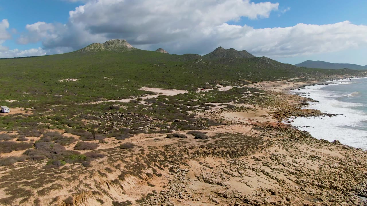 At Buzzards Beach East Cape BCS Mexico, an aerial frame shows sandy scrubland, a rocky shoreline, white surf at right, green hills in the background, partly cloudy sky, and no visible people.