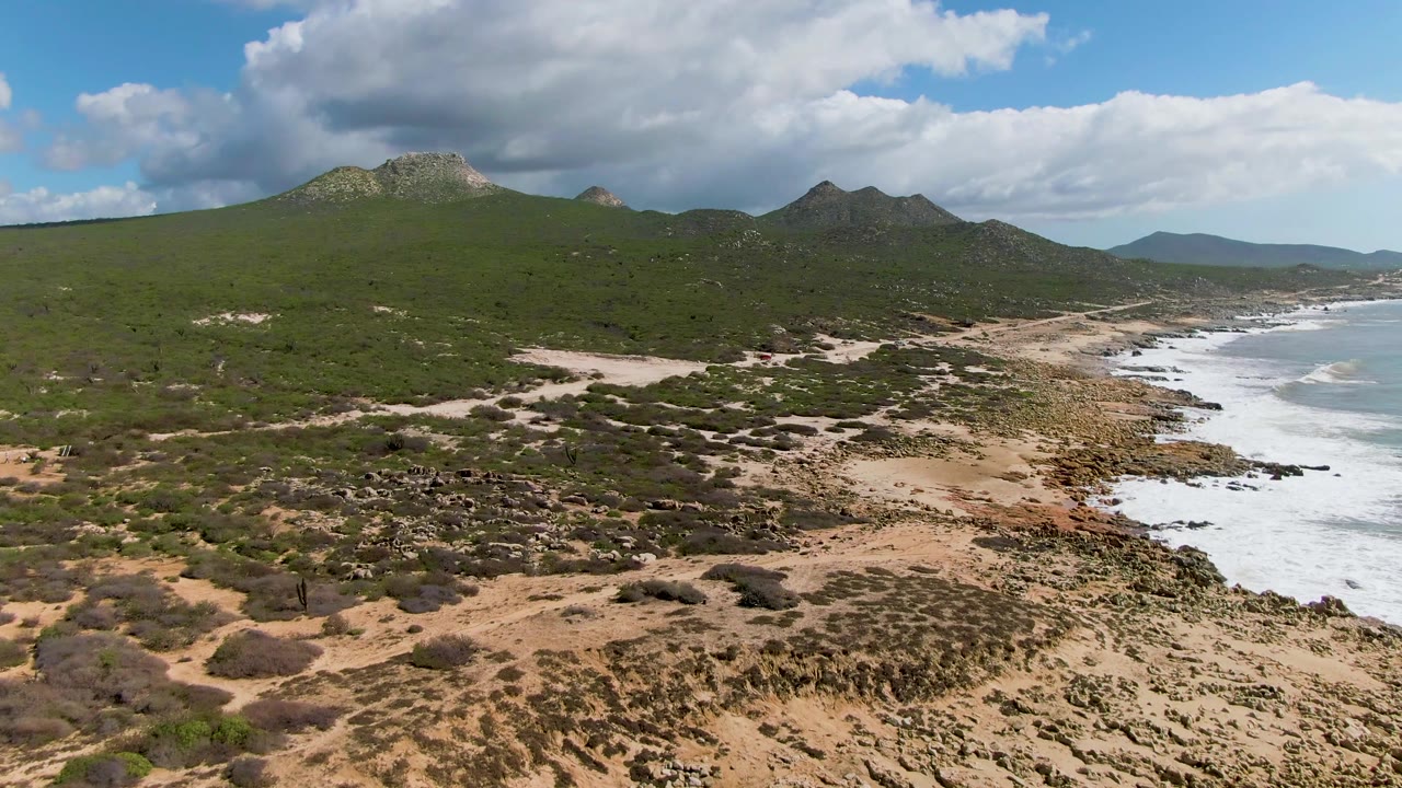 At 0:01, the Buzzards Beach aerial view centers more scrub-covered sand and dirt tracks while the rocky surf line runs along the right edge beneath green hills, with no visible people.