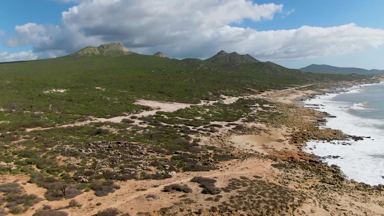 At 0:02, the aerial composition shows brushy dunes and scrub in the foreground, a curving rocky beach with white surf on the right, low mountains behind, and no visible people.