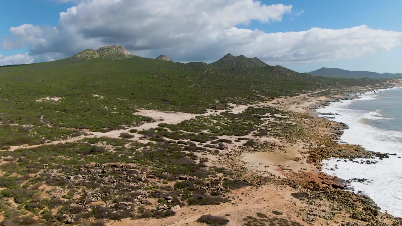 Aerial coastline with dark scrub patches, pale sand, rocky shore, surf, and hills.