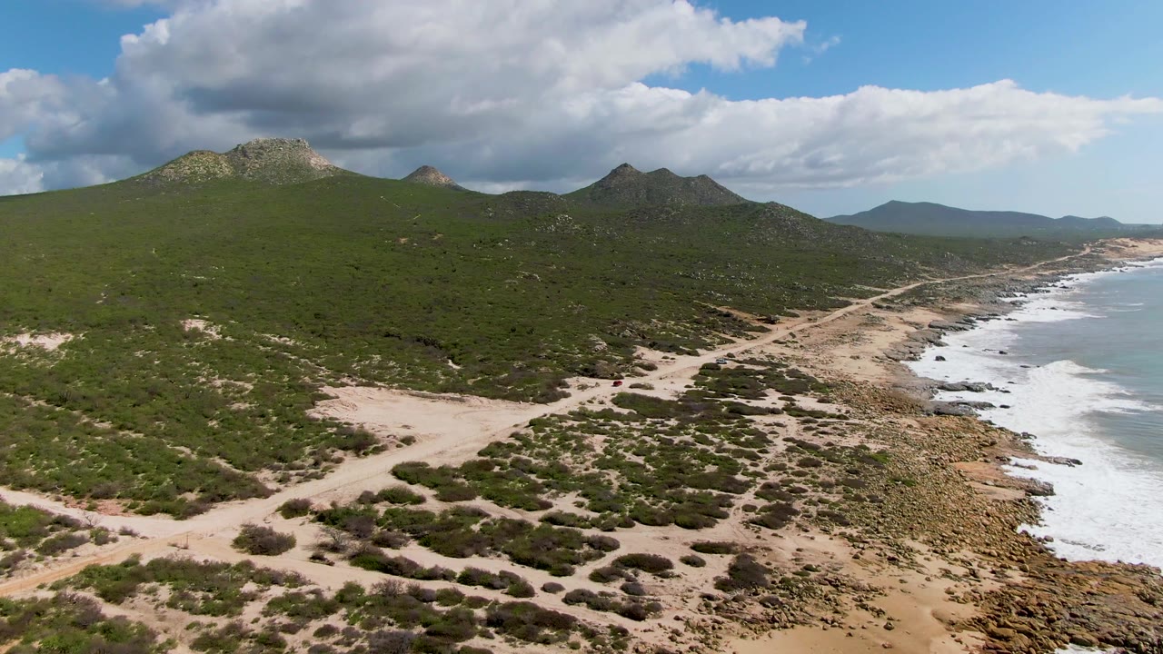 Aerial frame at Buzzards Beach East Cape BCS Mexico showing more of the sandy path network through low vegetation, with rocky shore and white-capped waves on the right and no visible people.