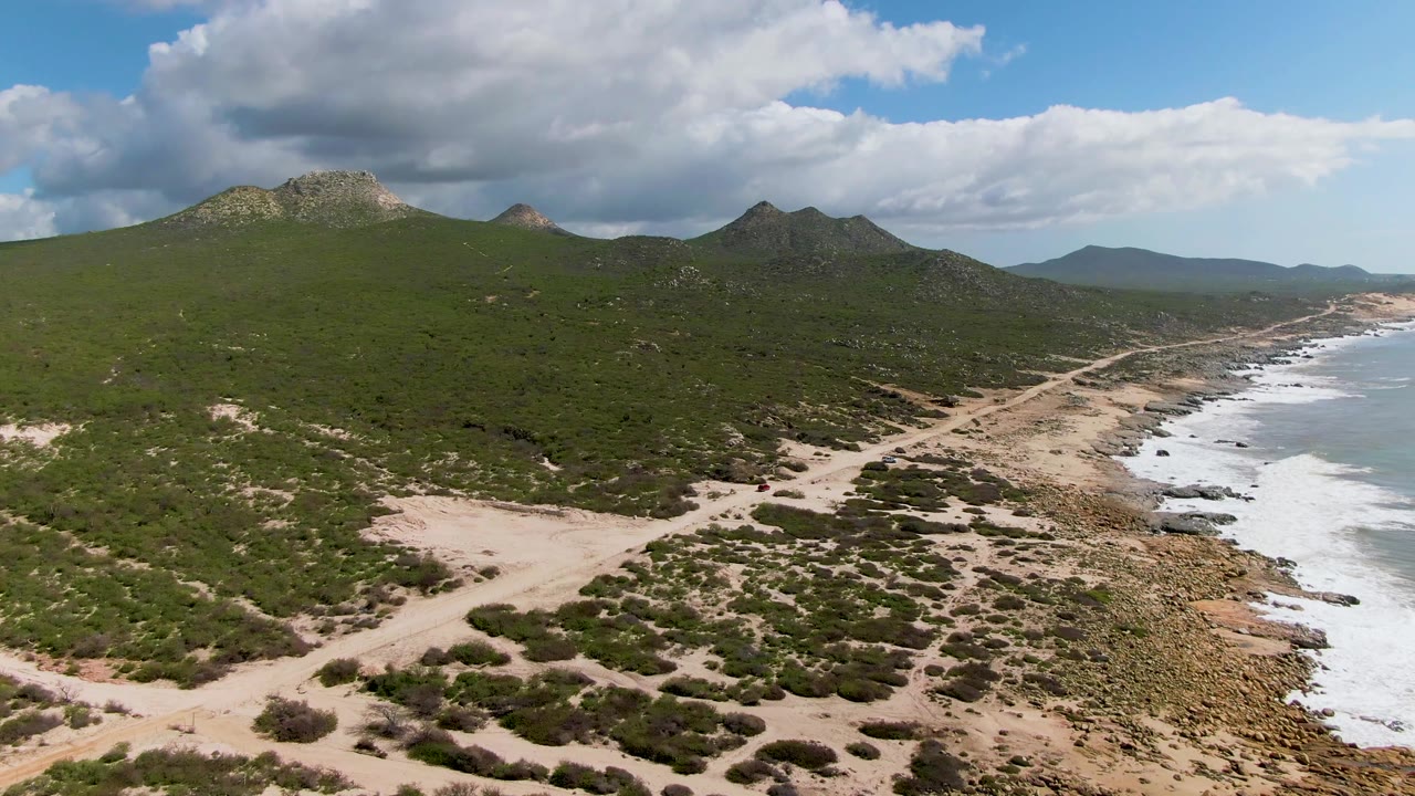 Mid-clip aerial view at Buzzards Beach East Cape BCS Mexico, with branching sandy tracks across scrub, rocky surf below the right side, distant hills on the horizon, and no visible people.