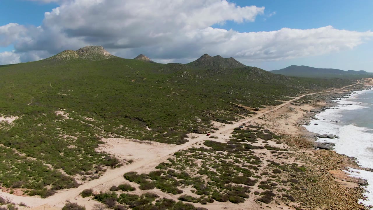 Aerial coastline view with sandy foreground, green mountain slopes, rocks, surf, and clouds.