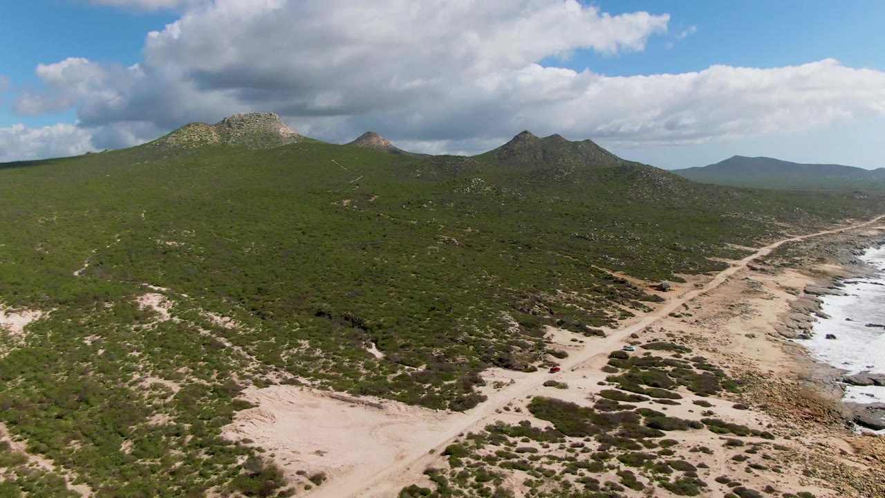 Aerial frame at Buzzards Beach East Cape BCS Mexico showing a sandy road near rocky surf, green hills under large clouds, a small red car near the lower right, and no visible people.