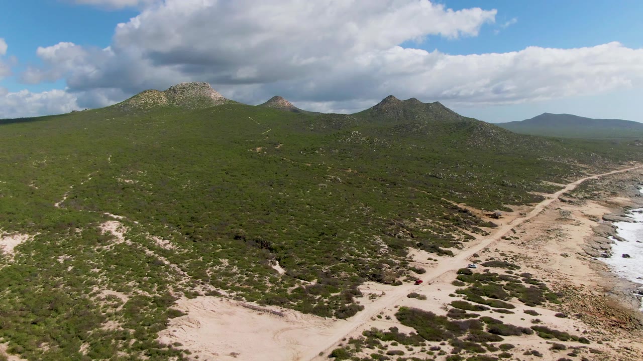 Aerial frame at Buzzards Beach East Cape BCS Mexico with the sandy path curving through scrub, the red car lower right, rocky shoreline at frame edge, distant hills, and no visible people.