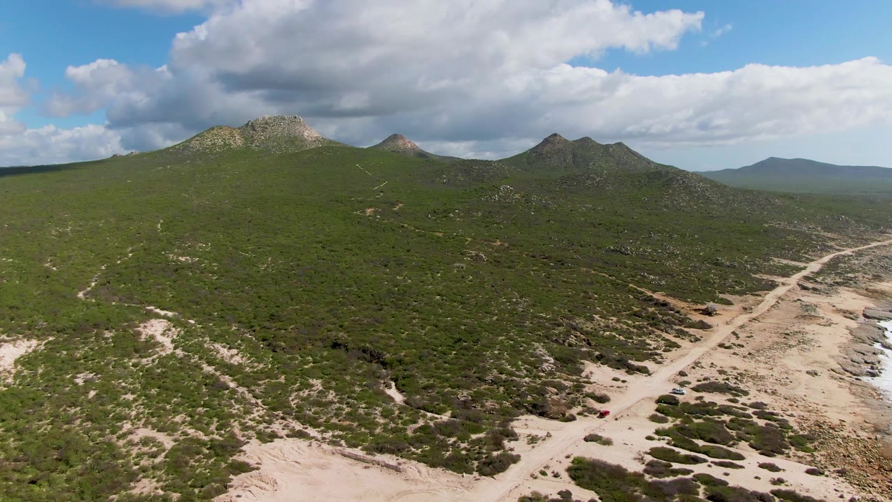 Aerial frame at Buzzards Beach East Cape BCS Mexico looking across green hills and a pale sandy road toward rocky surf, with the red car near the lower right and no visible people.