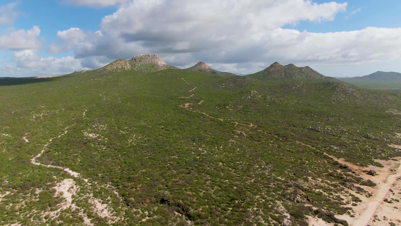 Aerial view in Buzzards Beach East Cape BCS Mexico with a small vehicle on the dirt road, green scrub covering the hills, rocky ridges beyond, and no visible people.