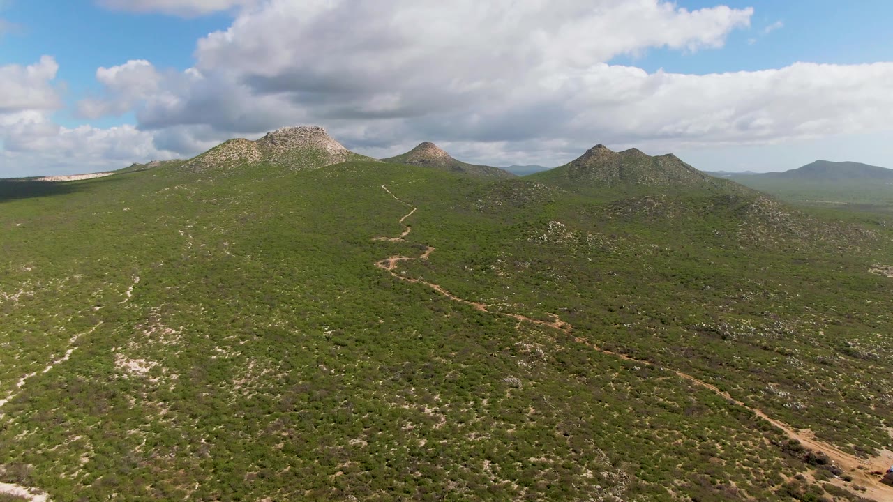 Aerial frame from Buzzards Beach East Cape BCS Mexico showing green scrub hills, a winding dirt road, rocky peaks on the horizon, partly cloudy sky, and no visible people.