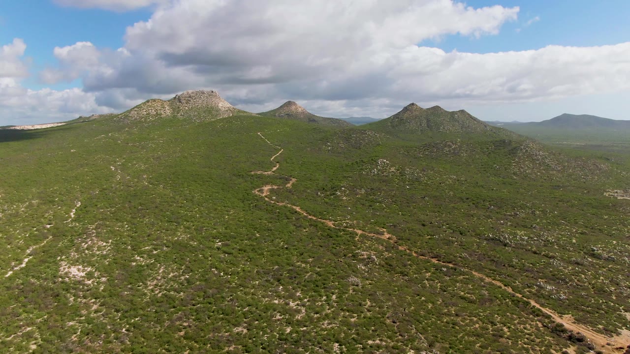 Second aerial frame from Buzzards Beach East Cape BCS Mexico with the dirt road more centered across scrub-covered hills, low rocky mountains in the background, cloud shadows, and no visible people.