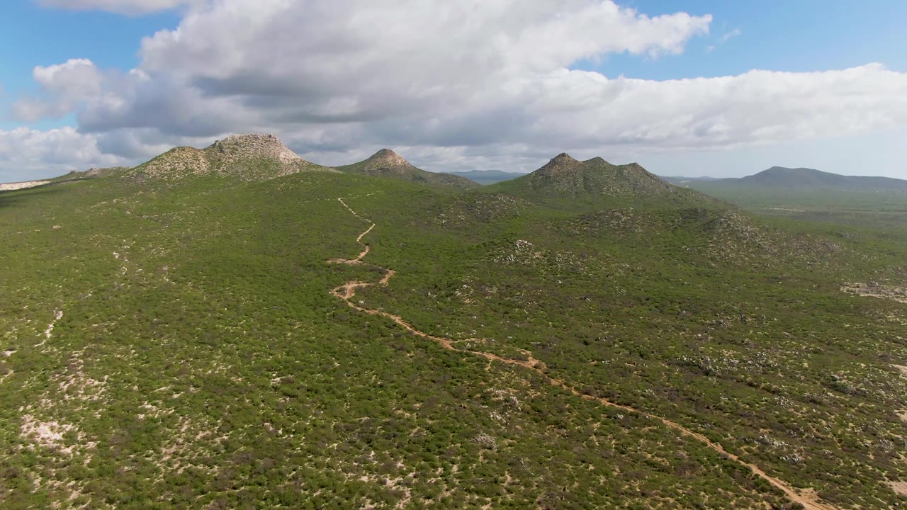 Fourth frame from Buzzards Beach East Cape BCS Mexico with the curving dirt road low in the frame, rounded green hills and rocky peaks ahead, broad clouds overhead, and no visible people.