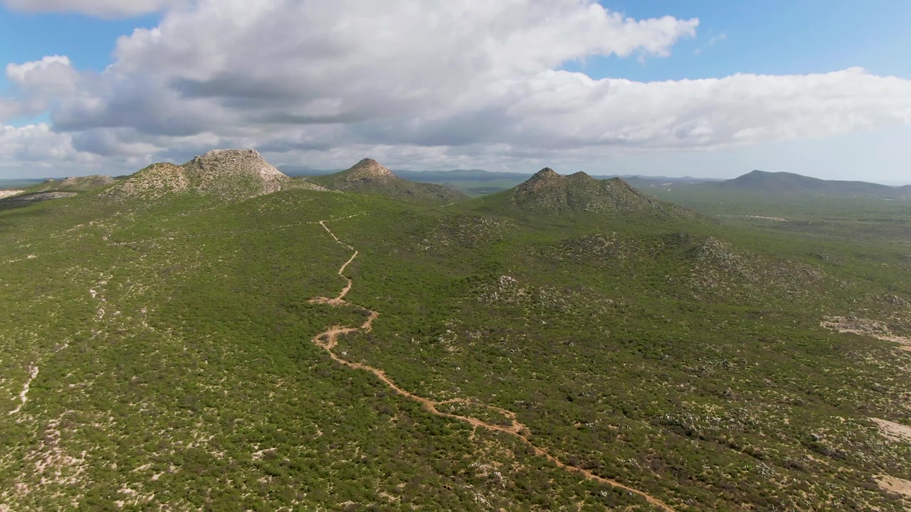 Aerial frame over the Mexico hillside, showing the dirt path leading up the green slope while cloud shadow darkens part of the valley and no visible people appear.