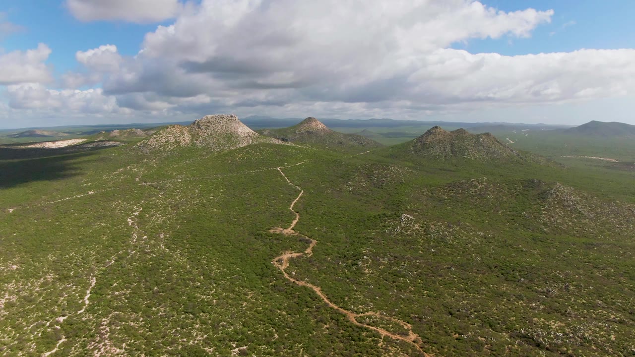 Wide aerial frame in Buzzards Beach East Cape BCS Mexico showing a dirt road through green mountain terrain, rocky hilltops in the background, and no visible people.