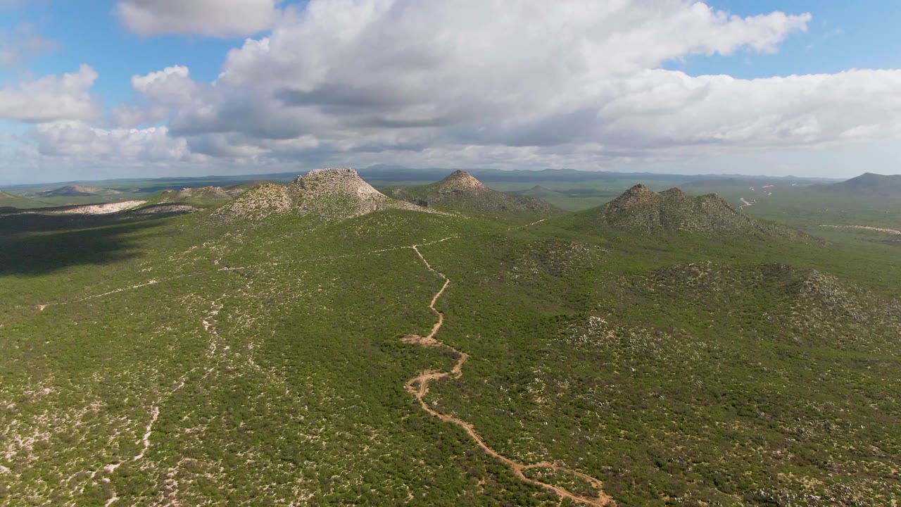 Aerial view of green hills with a winding dirt path and rocky ridges under scattered clouds.