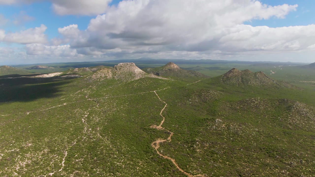 Opening aerial frame over Buzzards Beach East Cape BCS Mexico terrain, showing green hills, a winding dirt path, rocky ridges, distant horizon, and no visible people.