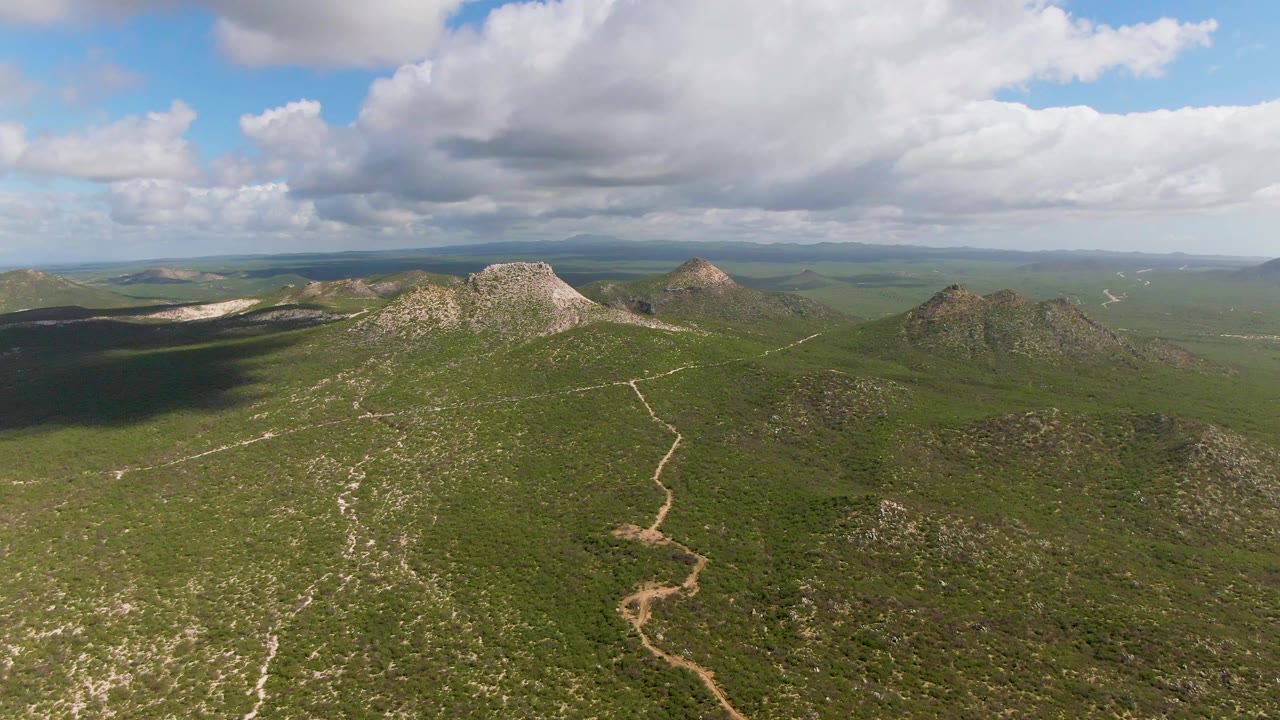 Green hillside aerial with rocky peaks, a winding trail, and cloud shadows.