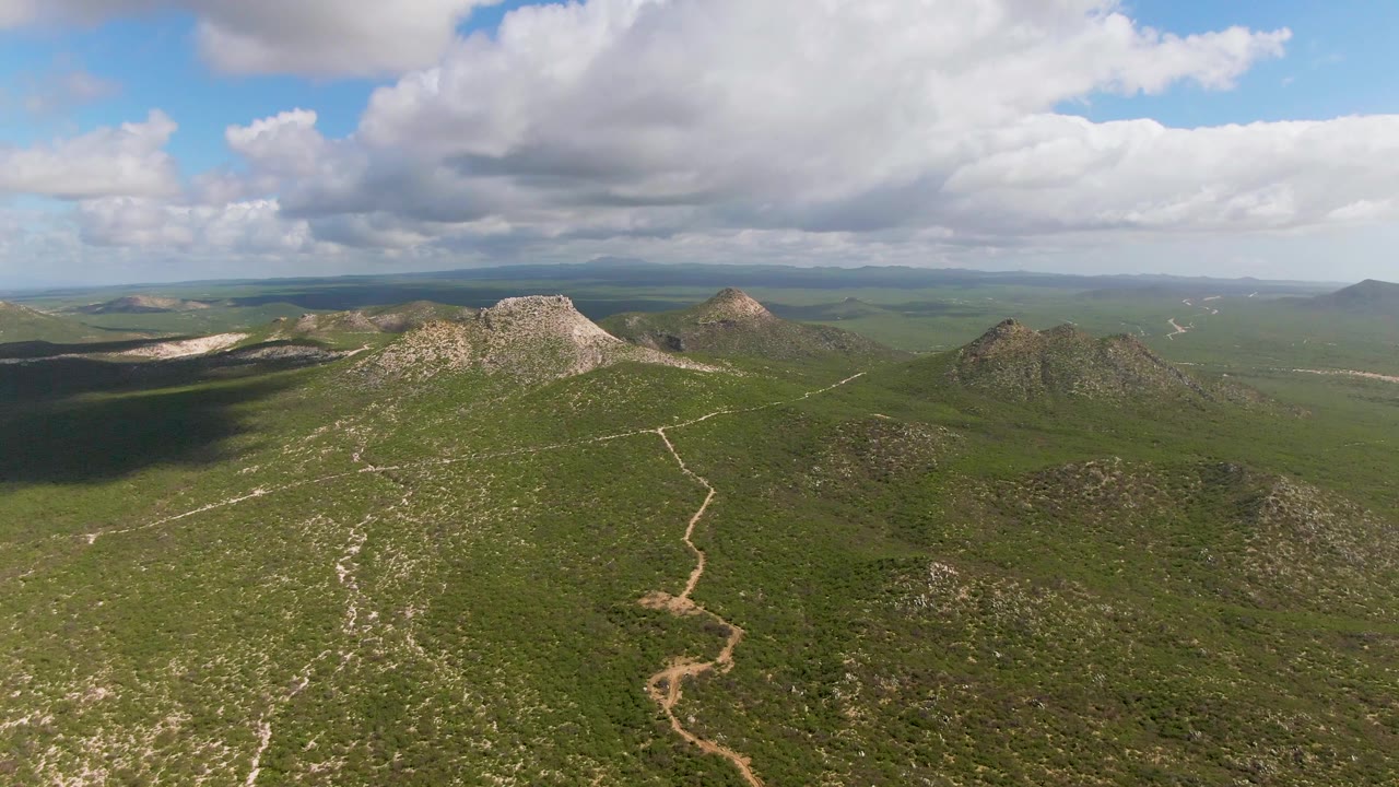 Aerial frame over Buzzards Beach East Cape BCS Mexico terrain, with green hilltops, a winding dirt road, distant horizon, broad cloud cover, and no visible people.