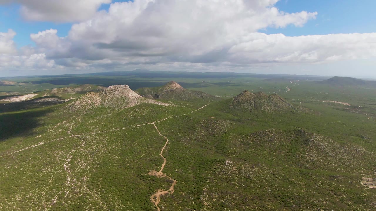 Aerial frame over Buzzards Beach East Cape BCS Mexico with a pale dirt trail running through scrub-covered hills, rocky outcrops, distant plains, cloud shadow, and no visible people.