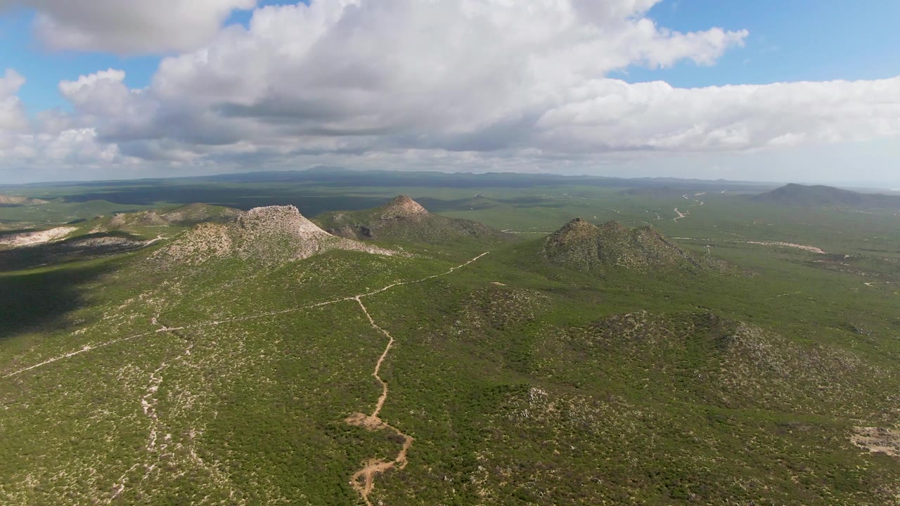 Wide aerial view in Buzzards Beach East Cape BCS Mexico showing clustered rocky hills in the foreground and midground, an open green valley beyond, broken cloud cover, and no visible people.