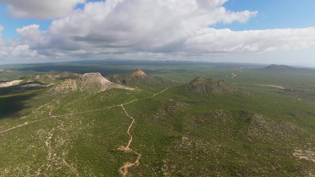Mid-clip aerial frame from Buzzards Beach East Cape BCS Mexico with rocky green hills, pale footpaths or dirt tracks, a darker cloud shadow at left, distant open land, and no visible people.