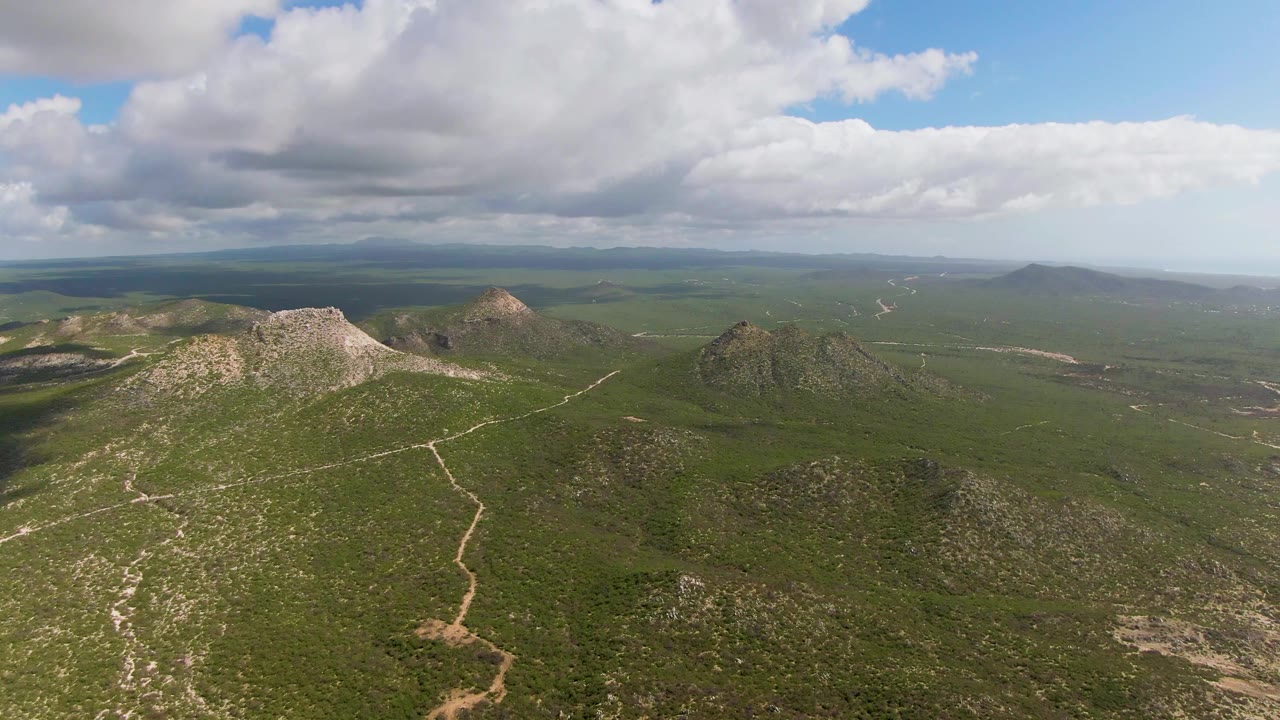 Wide drone view of Buzzards Beach East Cape BCS Mexico hills with scrubby green cover, pale dirt paths, rocky ridges, distant valleys, shifting cloud shade, and no visible people.