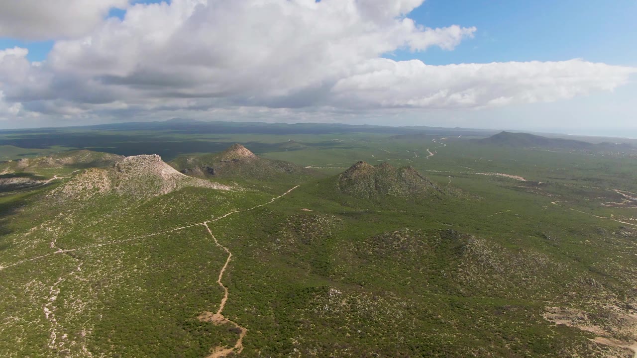 Aerial frame over Buzzards Beach East Cape BCS Mexico showing a central dirt trail crossing green hilly terrain, rocky peaks on both sides, layered distant land, and no visible people.