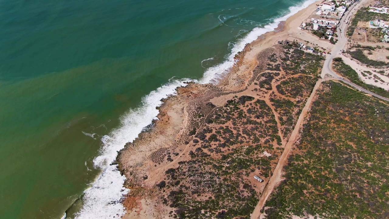 Aerial frame over Buzzards Beach East Cape BCS Mexico showing green water, white surf, a sandy rocky shore, scrub vegetation, dirt tracks, distant buildings, and no visible people.
